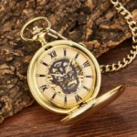 Steampunk skull mechanical pocket watch front view with exposed skeleton movement, Roman numeral dial, and gold-toned polished metal case, displayed on wooden surface with natural bark backdrop