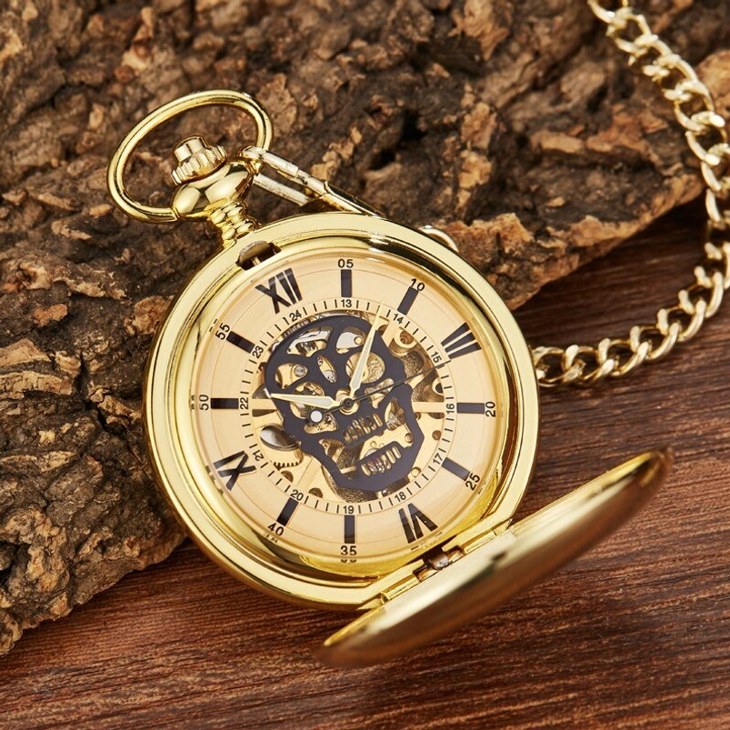 Steampunk skull mechanical pocket watch front view with exposed skeleton movement, Roman numeral dial, and gold-toned polished metal case, displayed on wooden surface with natural bark backdrop