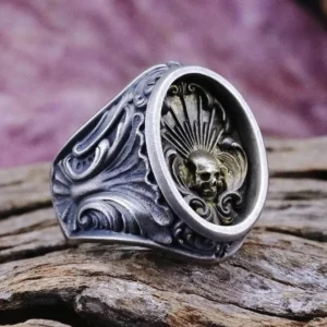 Steampunk skull ring with embossed design and ornate scrollwork band in vintage silver tone, displayed on weathered wood surface with purple fabric backdrop showing three-quarter side view