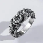 Oxidized silver steampunk ring with double-headed snake and skull design, three-quarter side view on minimalist white background, showing detailed gothic embossed motifs and weathered metallic finish