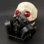 Steampunk spike gas mask with black plastic body, bronze metal chains, and cross-skull badge displayed on skull mannequin - front-left angled view showing adjustable elastic strap