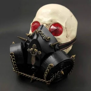 Steampunk spike gas mask with black plastic body, bronze metal chains, and cross-skull badge displayed on skull mannequin - front-left angled view showing adjustable elastic strap