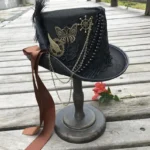 Steampunk top hat displayed on wooden stand, three-quarter left view showcasing black felt, bronze ornaments, brown satin ribbon and black feather details in outdoor setting
