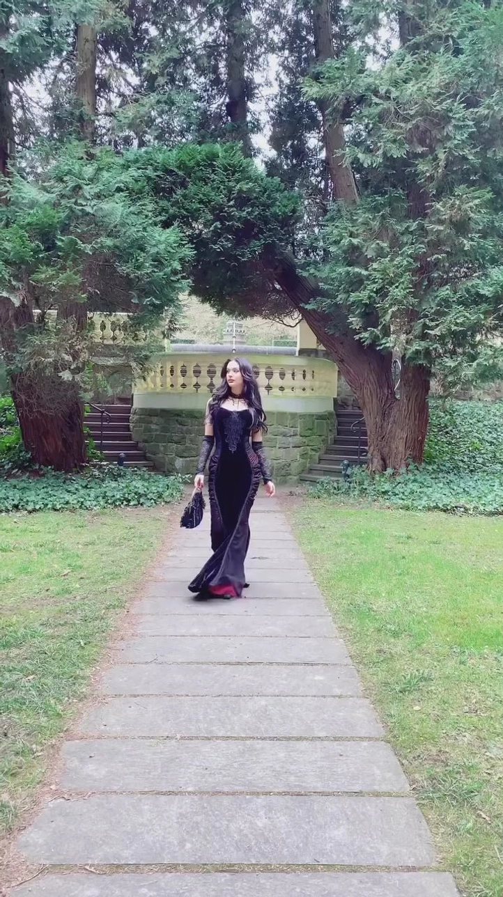Black red Victorian steampunk mermaid dress full distant view, complete outfit with mini handbag, long dark hair model, garden stone path with vintage architecture background