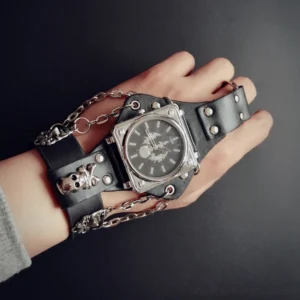 Close-up overhead view of steampunk square case watch featuring silver skull ornaments, black leather strap with snap buttons, and chain embellishments on dark background