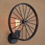 Steampunk spoked wheel wall lamp with lit Edison filament bulb, matte black metal frame mounted on white wall for home interior decor