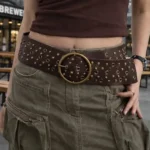 Female model wearing dark brown steampunk wide waist belt with antique brass round buckle and metal studs, outdoor cafe street setting, front view showcasing full belt aesthetic