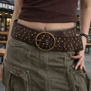 Female model wearing dark brown steampunk wide waist belt with antique brass round buckle and metal studs, outdoor cafe street setting, front view showcasing full belt aesthetic