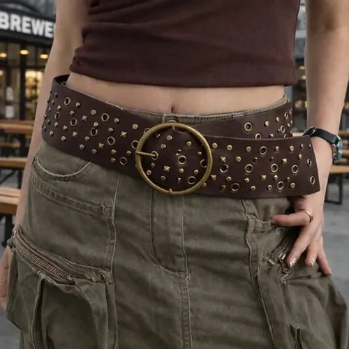 Female model wearing dark brown steampunk wide waist belt with antique brass round buckle and metal studs, outdoor cafe street setting, front view showcasing full belt aesthetic
