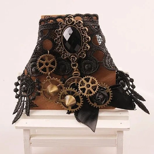 Frontal 3/4 view of black steampunk lace wrist cuff featuring intricate lace pattern, bronze gears, large black gem, and elegant satin bow detail on white wood surface