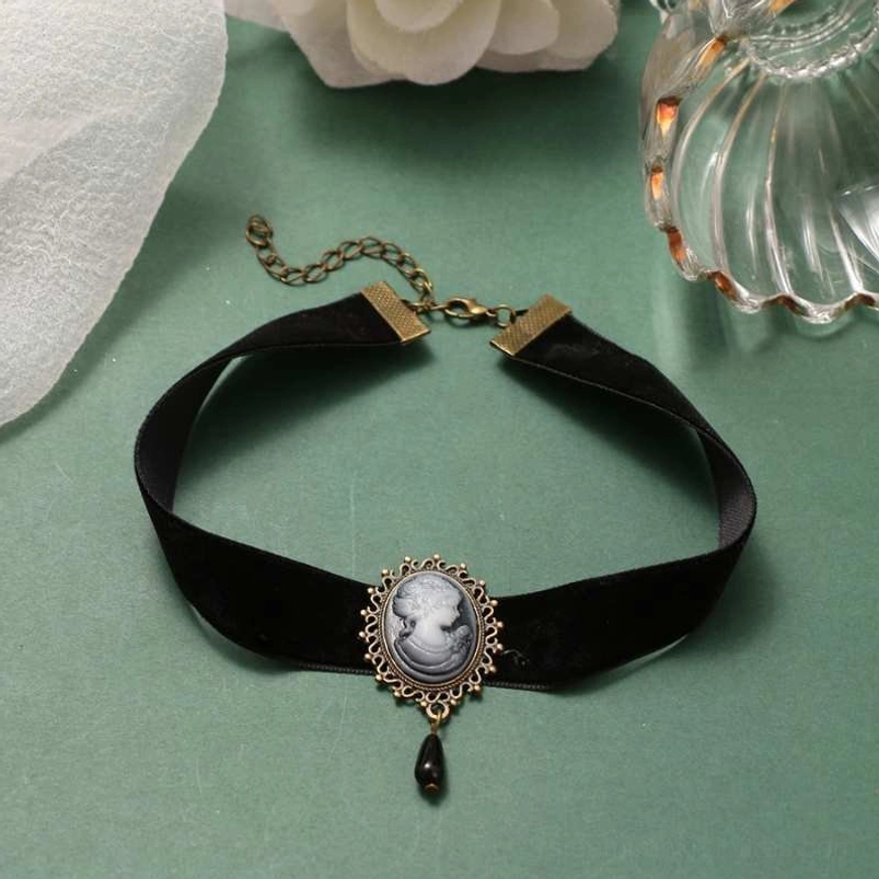 Unworn Victorian cameo velvet choker displayed flat lay on green tabletop with decorative props, featuring adjustable extension chain and bronze hardware details