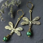 Vintage antiqued bronze dragonfly dangle earrings with iridescent green pearl pendants on dark matte background with dried flowers, showing intricate carved dragonfly wing details