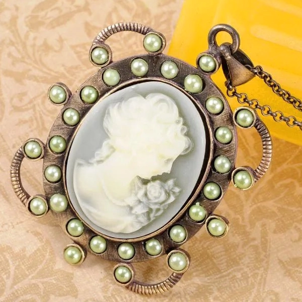 Close-up overhead view of hand-crafted Victorian cameo pendant with twisted bronze accents and full green pearl border, showing intricate carving details on damask textile