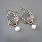 Pair of vintage pearl drop earrings with antiqued brass filigree and carved floral motifs, displayed in flat lay on gray background