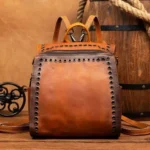 Vintage steampunk leather backpack front view featuring two-tone brown full-grain cowhide, metal grommet rivets, brass hardware buckles, and top zipper flap with industrial Victorian aesthetic on rustic wooden background