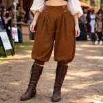 Women's steampunk pirate breeches front view, brown suede fabric with pleated front, buttoned waist, and cropped gathered leg cuffs, worn by female model outdoors at renaissance fair