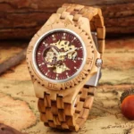 Light brown zebrawood steampunk watch with red skeleton dial, gold exposed movement, front three-quarter view on vintage map backdrop